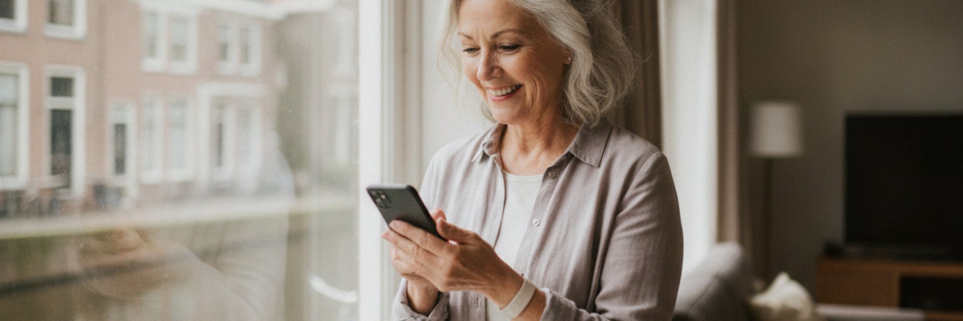 Vrouw bij het raam leest glimlachend een bericht op haar telefoon